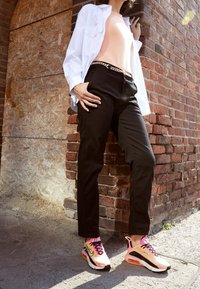 Svarta cargobyxor, ljus persikofärgad topp, vit skjorta med knappar och rosa-svarta sneakers med luftkuddar och kontrasterande snören, stående mot en tegelvägg.