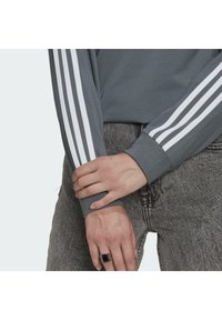 Grå långärmad skjorta med vita ränder på ärmarna, ihop med grå texturerade jeans. Noterbar ring på handen. Avslappnad passform och stil.