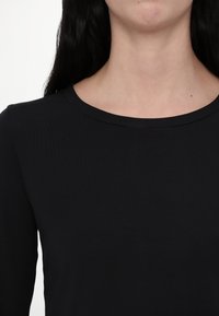 Femme portant un t-shirt noir uni à manches longues et col rond, montrant le cou et le haut de la poitrine avec de longs cheveux foncés.