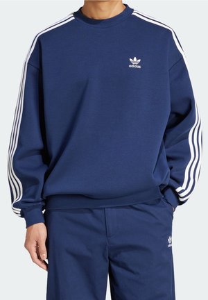 Mężczyzna ubrany w granatowy dres Adidas z białymi paskami na rękawach i spodniach, logo Adidas na piersi i udzie.