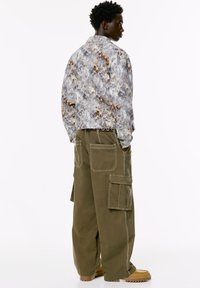 Uomo che indossa una giacca a fantasia grigia e marrone con pantaloni cargo olive verdi oversize e stivaletti color sabbia, in piedi di lato con le mani nelle tasche.