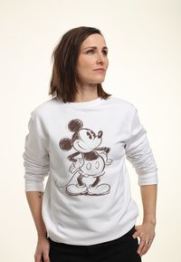 Felpa bianca con un disegno marrone di Topolino in posizione eretta. Il tessuto sembra morbido, con polsini a coste e una vestibilità rilassata.