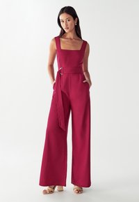 Ženska stoji z rokami v žepih, oblečena v brezrokavni rdeč kombinezon s širokimi hlačami in pasom ter bež sandali s peto na beli ozadju.