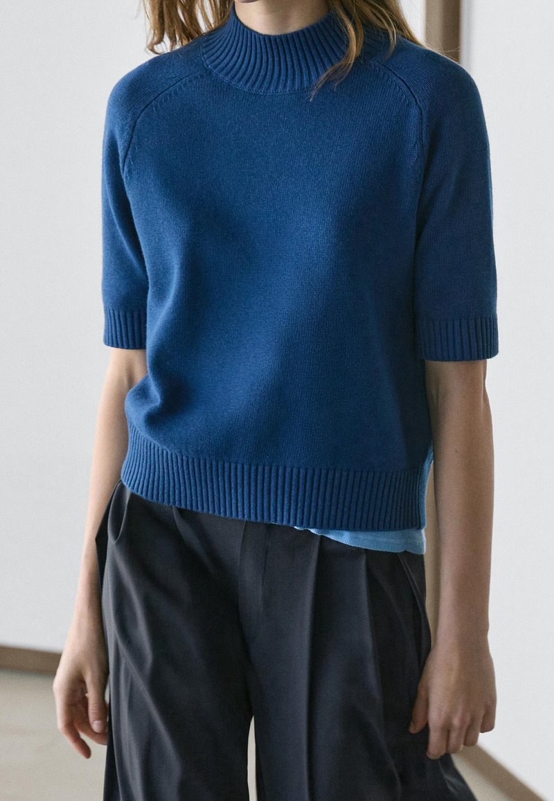 Femme portant un pull bleu en maille à manches courtes avec col, poignets et ourlet côtelés, assorti à un pantalon noir ample.