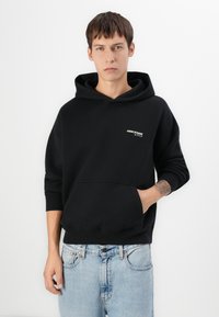 Sweat-shirt à capuche noir avec poche kangourou, poignets côtelés et logo brodé sur la poitrine, porté avec un jean denim bleu clair.