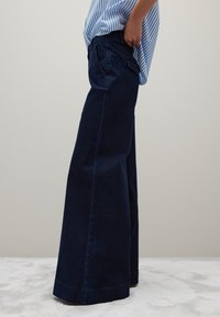 Pantaloni in denim a gamba larga di colore blu scuro, con vita alta, tasche laterali e una texture liscia. Abbinati a una maglietta a righe non inserita.