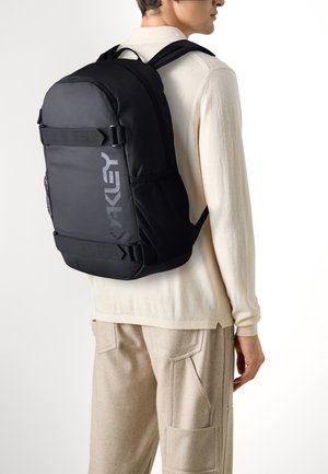 Un sac à dos noir en nylon avec des bretelles réglables, un grand compartiment principal, des poches latérales en maille et un logo proéminent à l'avant.