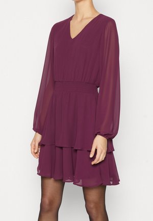 Femme portant une robe à manches longues, de couleur violet profond, à col en V, avec une taille cintrée et une jupe à volants, assortie à des collants noirs transparents.