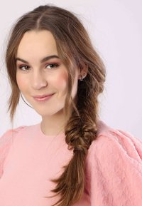 Jonge vrouw met lang, golvend bruin haar in een losse vlecht, gekleed in een lichtroze gemêleerde top met pofmouwen. Neutrale achtergrond.