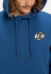 Blue hoodie made from soft fabric featuring a logo patch on the chest, adjustable drawstrings, and a relaxed fit.