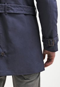 Navy sininen trench-takki, jossa on vyöllinen vyötärö, sileä pinta, nappiyksityiskohtia sekä räätälöity istuvuus hihansuissa ja helman kohdalla.