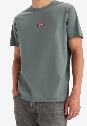 Hombre con camiseta verde de Levi's de manga corta y vaqueros grises, con las manos en los bolsillos, mirando hacia adelante.