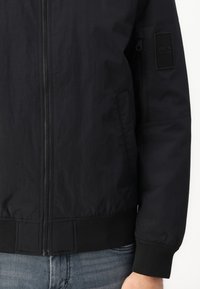 Mustmust must jacket, mis on valmistatud kergest materjalist, esifliisi, ribikätega ja tekstuuriga plaastriga vasakul varrukal.