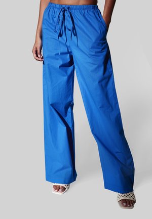 Blå bukser med vide ben og snøre i taljen, båret sammen med hvide åbne flettede hæle, kun den nedre del af overkroppen og benene er synlige.