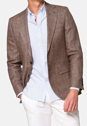 Mann trägt einen braun karierten Blazer über einem hellblau gestreiften Hemd mit weißen Hosen, posiert mit einer Hand in der Tasche.
