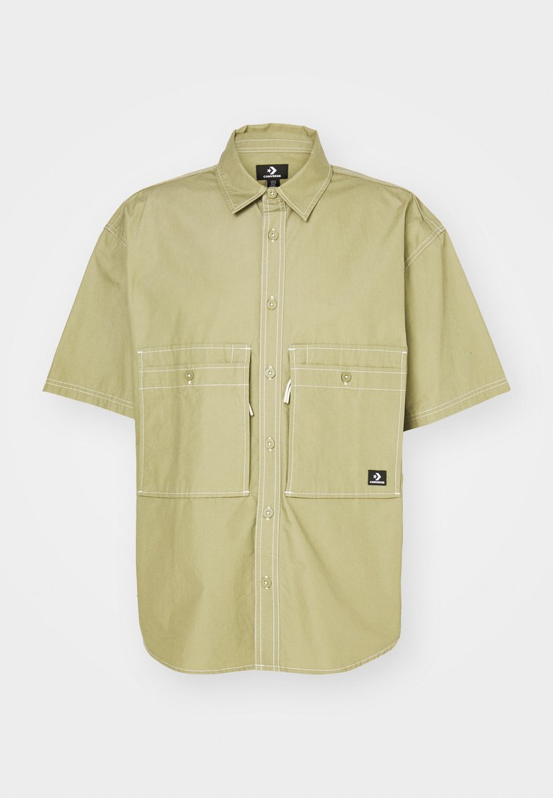 Camisa de manga corta en algodón verde oliva claro con costuras blancas, dos grandes bolsillos en el pecho y un pequeño logo de la marca en la parte inferior derecha.