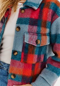 Károvaná bunda v modrej, červenej a tyrkysovej vlne. Obsahuje dve predné vrecká, hnedé gombíky a golier. Mäkka textúra.