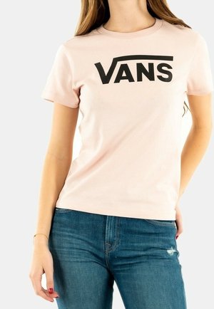 Kobieta w jasnoróżowej koszulce z krótkim rękawem i dużym czarnym logo „VANS”, w połączeniu z niebieskimi dżinsami z lekkim przetarciem.