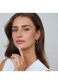 Guldtonade örhängen med en vågig design och slät yta. Ämnet har långt brunt hår, neutral sminkning och en vit klänning med V-ringning.