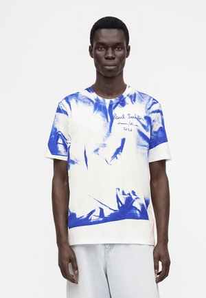 Homme portant un t-shirt tie-dye blanc et bleu avec le texte "Paul Smith Autumn Winter 2026" sur la poitrine, assorti à un pantalon de couleur claire.