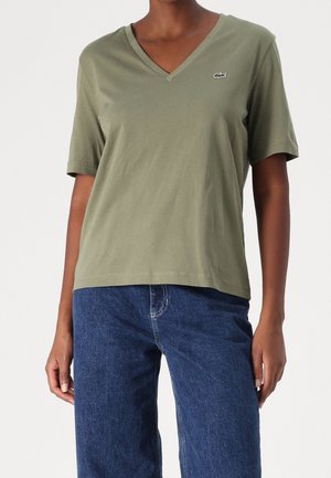 Personne portant un T-shirt vert olive à col en V et manches courtes avec un petit logo sur la poitrine et un jean denim bleu, debout devant un fond blanc.