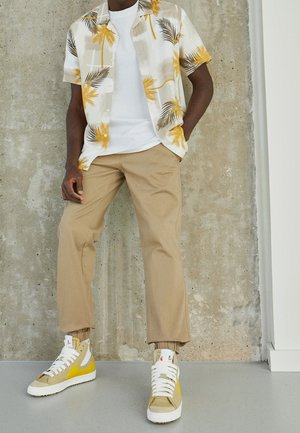 Homme portant un pantalon beige, un T-shirt blanc, une chemise à manches courtes avec un imprimé de palmier jaune et des baskets montantes beige et jaune.