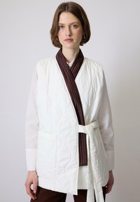 Vatteret vest i off-white, med en mørkebrun, ribbet sjalskrave, bælte i taljen og to frontlommer. Glat, blød tekstur.