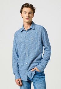 Camicia button-up a quadri blu e bianchi con taschino sul petto, realizzata in tessuto con una texture liscia. Presenta maniche lunghe e colletto.