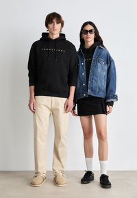 Sudadera negra con capucha y logo dorado, pantalones beige y zapatos slip-on color tostado en un hombre. Mujer con chaqueta de mezclilla azul, camiseta negra y falda negra.