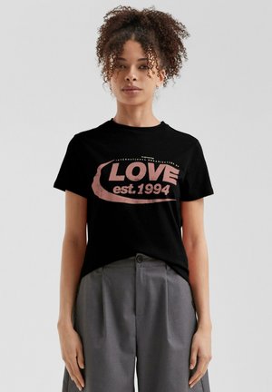 Schwarzes Baumwoll-T-Shirt mit einem pinken Grafikdruck, der "LOVE est. 1994" zeigt. Kombiniert mit grauen, hochgeschnittenen Hosen.