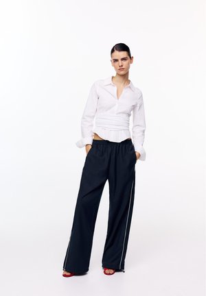 Femeie cu părul dat pe spate, purtând o cămașă albă mulată, pantaloni negri largi cu dungi laterale, pantofi roșii decupați în față, mâinile în buzunare, fundal neutru.