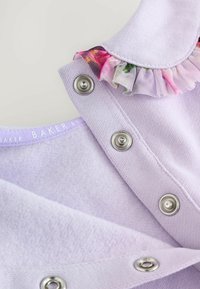 Vêtement en coton lavande avec un col à volants orné de fleurs, boutons-pression argentés et une finition douce et texturée.