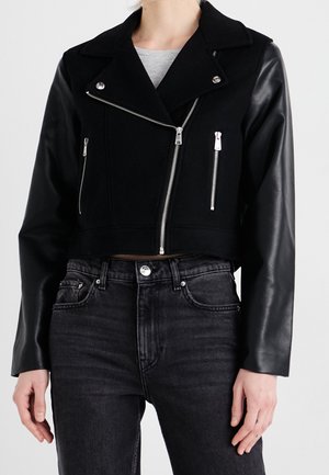 Black cropped jacket with leather sleeves and silver zippers worn over gray top with high-waisted black jeans on a standing person.