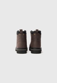 Un par de botines de cuero marrón con cuellos acolchados negros y suelas gruesas negras, mostrados desde la parte posterior sobre un fondo blanco liso.