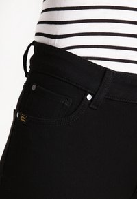 Jeans de mezclilla negra de corte slim, con un clásico diseño de cinco bolsillos. Incluyen herrajes plateados y una pequeña etiqueta de la marca. Camisa a rayas visible.