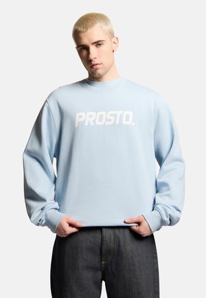 Młody mężczyzna z krótkimi blond włosami, noszący jasnoniebieską bluzę z napisem "PROSTO.", trzymający krawędź, ubrany w ciemne jeansy.