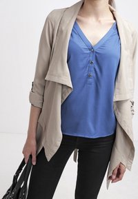 Beige lightweight jacket over a blue V-neck top with buttons, paired with black pants. Model holding a black handbag, plain white background.