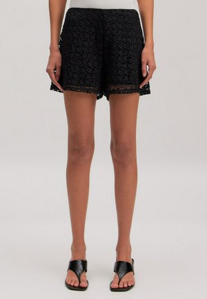 Vrouw die zwarte kanten shorts en zwarte sandalen met open tenen draagt, staat met ontspannen handen langs haar zij op een lichte achtergrond.
