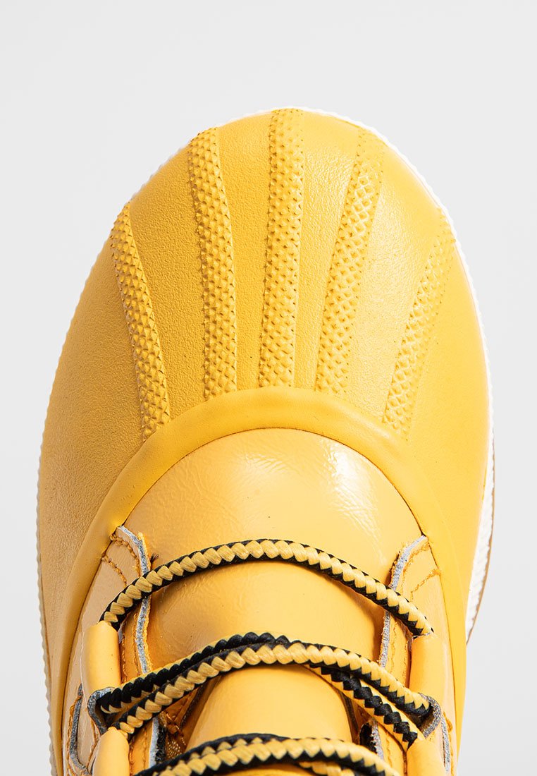 Żółty gumowy but z teksturowanym noskiem, wypukłymi paskami i czarnymi sznurówkami. Gładkie wykończenie, zaokrąglony kształt i biała gumowa podeszwa.