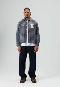 Blauw en grijs geruit overhemd met knopen en een witte geborduurde letter "E", gedragen over een wit shirt, gecombineerd met donkere spijkerbroeken en bruine schoenen.