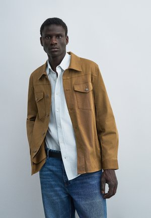 Homme portant une veste marron boutonnée par-dessus une chemise blanche et un jean bleu, debout devant un fond uni gris clair.