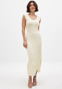 Strickkleid in Off-White mit figurbetonter Silhouette, runder Ausschnitt, geripptem Design und seitlichem Schlitz. Akzentuiert mit metallic Sandalen mit Riemchen.