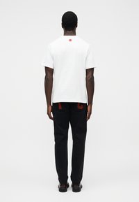 Λευκό κοντομάνικο μπλουζάκι με κόκκινο λογότυπο στο πίσω μέρος, συνδυασμένο με μαύρο παντελόνι με κόκκινα γράμματα στις τσέπες. Μαύρα παπούτσια.