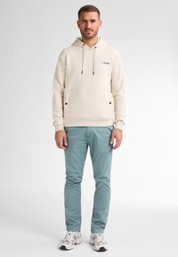 Helle cremefarbene Kapuzenjacke mit Fronttasche, kontrastierenden Zugbändern und Logo. Kombiniert mit hellblauen Hosen und weißen Sneakers.