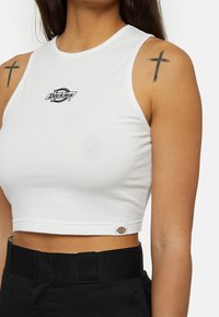 Witte katoenen cropped tanktop met een zwart "Dickies" logo op de borst en een merketiket aan de zoom. Zwarte high-waisted broek.