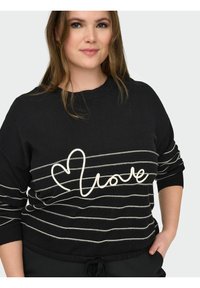Femme aux cheveux châtain clair portant un pull noir à rayures blanches horizontales avec un motif en forme de cœur et le mot "amour", main dans la poche.