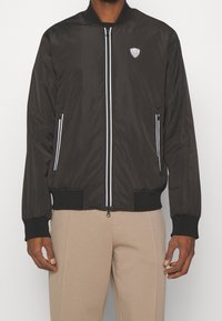 Svart bomberjacka av slät tyg, med vita dragkedjsdetaljer, rund krage och ribbade ärmmuddar. Logotyp på bröstet.