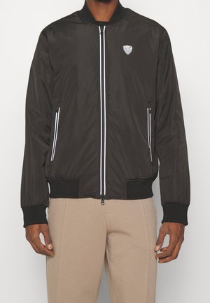 Chaqueta bomber negra hecha de tela suave, con detalles en blanco en el zipper, un cuello redondo y puños acanalados. Logotipo en el pecho.
