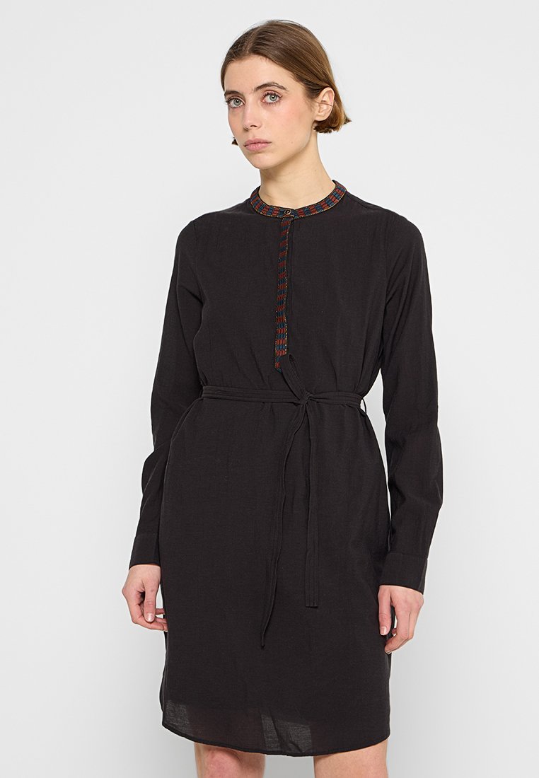 Schwarzes Langarmkleid mit Kragen, das einen bunten gewebten Besatz, eine zentrale Knopfleiste und einen Stoffgürtel in der Taille aufweist.