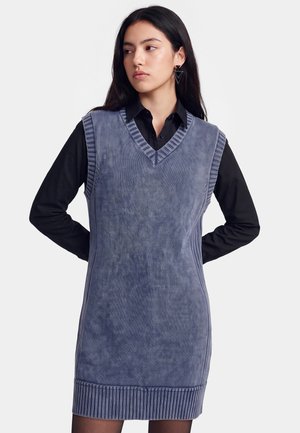 Junge Frau trägt ein blaues ärmelloses Strickkleid über einem schwarzen Langarmshirt und schaut seitlich mit den Händen hinter dem Rücken.